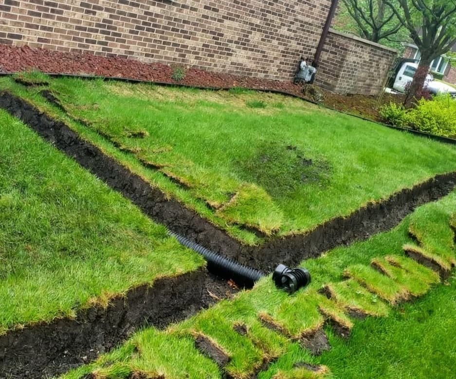 A trench cut through a front lawn with sod strips peeled back and corrugated drain pipe laid out